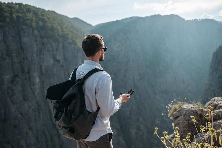 A man with a backpack and compass stands on a cliff, gazing at a scenic gorge.